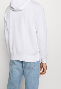 Person som bär en enkel vit hoodie och ljusblå denimjeans, sedd bakifrån mot en enkel vit bakgrund.