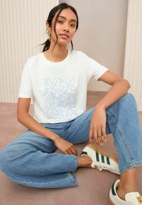 T-shirt blanc à manches courtes avec un motif floral bleu clair et le texte "keep growing" sur le devant. Associé à un jean bleu ample et des baskets blanches.