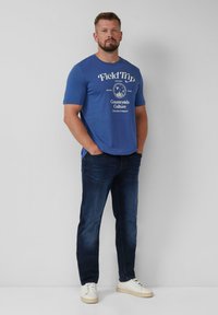 Blauwe T-shirt met korte mouwen, voorzien van witte tekst en graphics, gecombineerd met donkere jeans en witte sneakers. Casual pasvorm en een gladde stofstructuur.