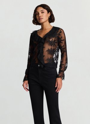 Camicia nera trasparente con ricami floreali in pizzo, dettaglio con balze sulla parte anteriore e maniche lunghe con polsini con bottoni, indossata con jeans neri.