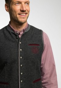 Dunkelgraue Wollweste mit silbernen Knöpfen, roten Samt-Akzenten und besticktem Logo; über einem rot-weißen karierten Hemd getragen.