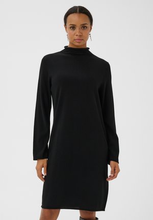 Femme aux cheveux foncés portant une robe noire à manches longues avec un col montant sur un fond clair uni.