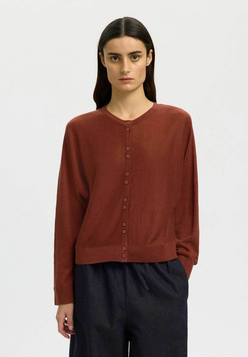 Femme aux longs cheveux foncés porte un cardigan boutonné couleur rouille et un pantalon ample foncé, debout devant un fond clair uni.