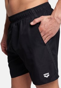 Bañador negro con cintura elástica, bolsillos laterales, costuras blancas en contraste y un logo blanco en la parte inferior delantera.