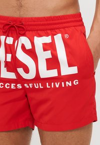 Červené plavky vyrobené z ľahkého materiálu, s veľkým bielym logom "DIESEL" a textom "SUCCESSFUL LIVING" v tučných písmenách. Nastaviteľná šnúrka na sťahovanie.