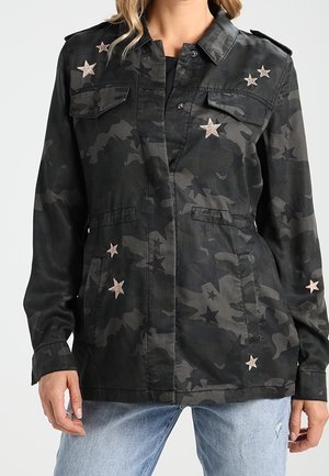 Black camo jacket with star patches on chest, sleeves, and pockets worn over black top with light blue jeans.
