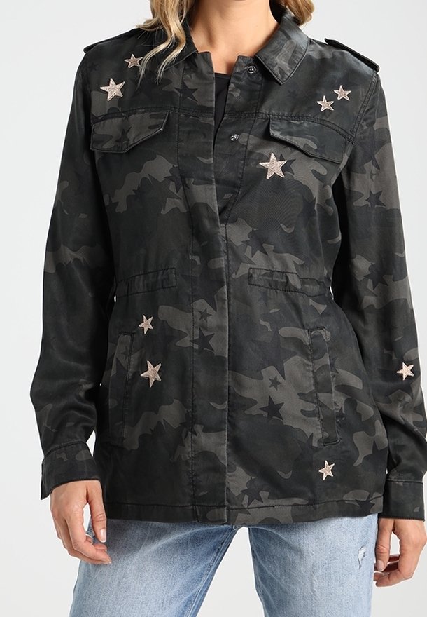 Veste camouflage noire avec des patchs étoiles sur la poitrine, les manches et les poches, portée sur un haut noir avec un jean bleu clair.