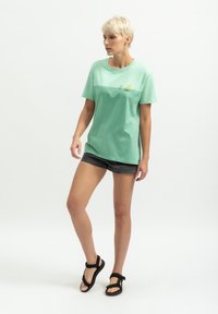 Lichtgroene katoenen t-shirt met korte mouwen en een relaxte snit, met een subtiele grafisch ontwerp aan de voorkant. Gecombineerd met donkere shorts.