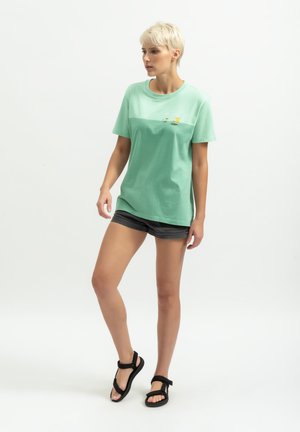 T-shirt en coton vert clair à manches courtes et coupe décontractée, avec un design graphique subtil sur le devant. Associé à des shorts foncés.