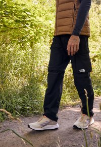 Svarta utomhusbyxor med cargofickor, bärs med en ljusbrun pufferväst och beige löparskor med lila ränder och orange detaljer.