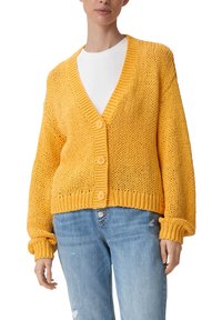 Keltainen neulecardigan, jossa on V-pääntie, napitus ja leveät hihat. Yhdistetty valkoisen paidan kanssa ja vaaleansiniset farkut.