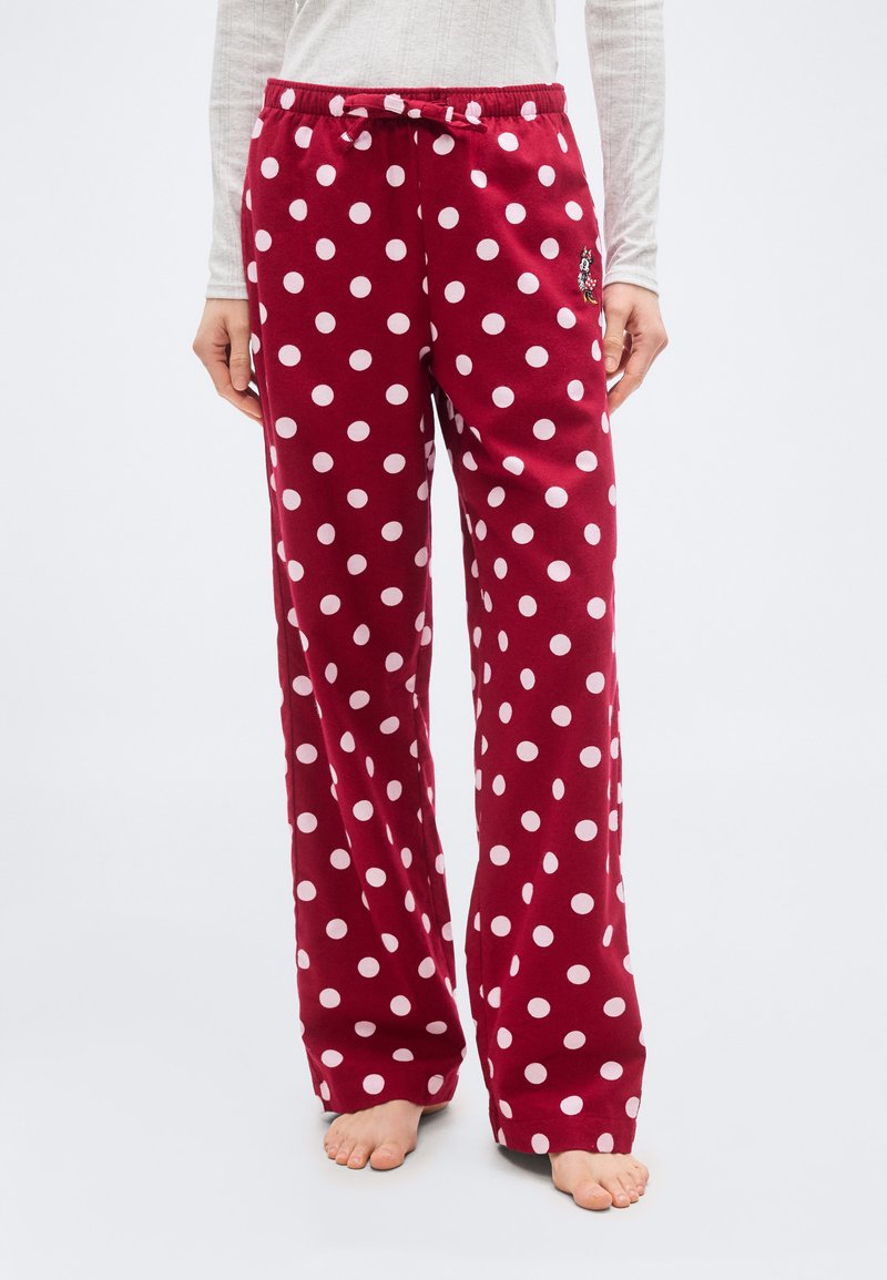 Άτομο που φοράει κόκκινο παντελόνι πιτζάμας με λευκές πουά και ένα μικρό καρτούν σχέδιο στη μεριά, που στέκεται ξυπόλυτο σε λευκό πάτωμα.