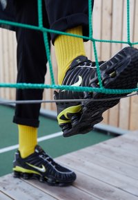 Personne portant des baskets Nike noires et jaunes et des chaussettes jaunes marchant sur un filet de corde vert au-dessus d'une plateforme en bois et d'un terrain de sport.