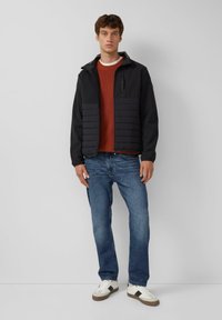 Zwarte gewatteerde jas met rits en hoge kraag over een oranje gebreid sweater. Gecombineerd met blauwe jeans en witte sneakers met bruine accenten.