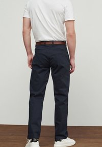 Man wearing dark navy trousers, white t-shirt, brown belt, and white sneakers, standing on wooden floor against plain light wall.
