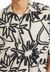 Košile s krátkým rukávem vyrobená z lehké tkaniny, s krémovým pozadím a výraznými černými květinovými vzory ve stylu štětce a malými knoflíky.