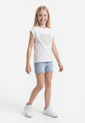 Χαμογελαστή νεαρή κοπέλα που φοράει λευκό αμάνικο μπλουζάκι με σχέδιο καρδιάς, ανοιχτόχρωμο τζιν σορτς και λευκά αθλητικά παπούτσια, στέκεται σε λευκό φόντο.