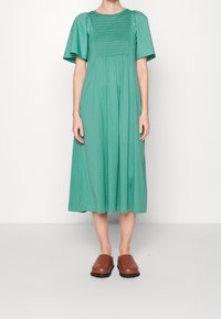 Robe midi verte avec des manches bouffantes courtes et un corsage smocké. Tissu lisse, coupe décontractée, portée avec des sabots marron.