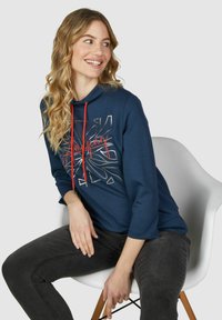 Marineblauwe hoodie met rode trekkoorden, voorzien van een zilveren grafisch ontwerp en driekwart mouwen. Gekleed met donkere jeans, zittend op een witte stoel.