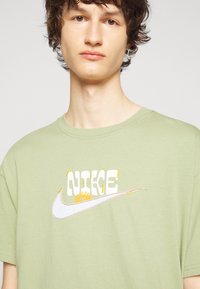 T-shirt en coton vert clair avec un logo Nike blanc brodé de accents jaunes et un graphique de soleil. Encolure ras du cou et coupe détendue.