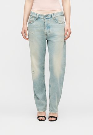 Donna che indossa jeans dritti color azzurro sbiadito con dettagli usurati e tacchi neri aperti sulle dita, in piedi davanti a uno sfondo bianco.