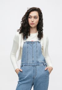 Lyseblå denim overaller med avslappet passform og justerbare stropper, kombinert med en kremfarget langermet topp med en liten rød grafikk.