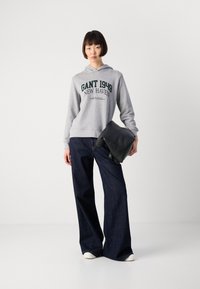 Jeune femme portant un sweat à capuche gris GANT 1949, un jean large noir, des baskets blanches, tenant un sac plié noir contre un fond blanc.