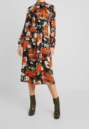 Kvinde iført en sort midi-kjole med orange og hvide blomsterprint, lange ærmer, talje med elastik, kombineret med mørkebrune ankelstøvler.