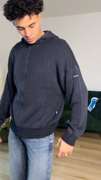 Svart huva med dragkedja, ribbad textur, långa ärmar och en figurnära midjeresår; bärs över blå jeans.