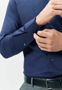 Sötétkék gombos ing, texturált anyagból, sötét gombokkal és állítható mandzsettákkal, fekete bő övvel párosítva.