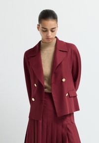 Young woman in burgundy double-breasted blazer with gold buttons over beige textured turtleneck and matching pleated skirt, looking down.