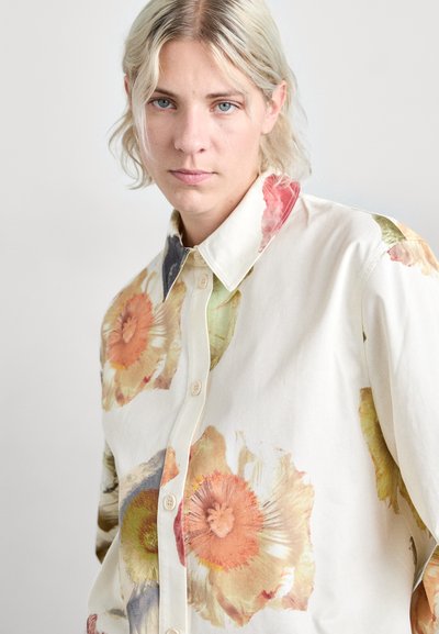 Camicia button-up con stampa floreale crema, caratterizzata da grandi fiori nei toni pastello, colletto a punta e tessuto testurizzato. Quattro bottoni sulla parte frontale.