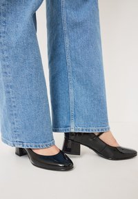 Black patent leather block-heeled shoes with a rounded toe, paired with light blue wide-leg jeans featuring a hemmed edge.