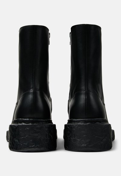Botas de tobillo de cuero negro con una suela gruesa y texturizada. El diseño presenta un superior suave con costuras visibles y un cierre de cremallera en el lateral.