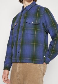 Blå och grön rutiga skjorta i texturerat tyg, med två framfickor och knäppning, med kontrasterande beige linjer i mönstret.