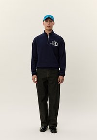 Jovem rapaz usando um boné azul, camisola azul-marinho com fecho de quarto de zíper e logótipo "LD", jeans pretos e sapatos pretos, em pé contra um fundo claro e simples.