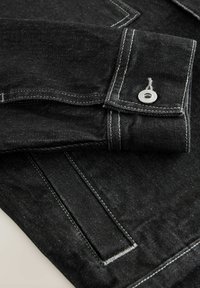 Dettagli di una giacca di jeans nera con cuciture bianche, polsini arrotolati e bottone a pressione argenteo. Texture e linee di cucitura visibili.