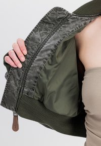 Maslinasto zelena bomber jakna s prešanim vanjskim dijelom, rebrastim manžetama i glatkom unutarnjom podlogom. Ima zatvarač i kožni trak.