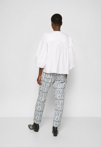 Blusa bianca con maniche a sbuffo e schiena tonda, abbinata a pantaloni tapered a stampa blu e bianca, indossati con scarpe con tacco a blocco nere.