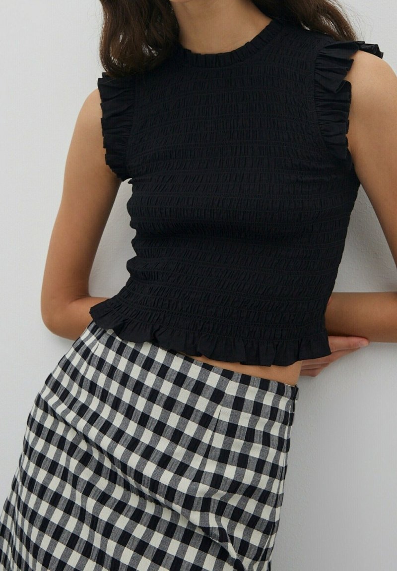 Femme portant un haut noir sans manches à volants et une jupe taille haute à carreaux noir et blanc, debout contre un mur clair uni.