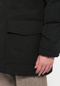 Casaco acolchoado preto com um grande bolso frontal, mangas estruturadas com textura acolchoada e fecho de botão seguro no punho.