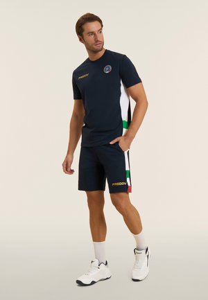 Uomo in piedi con abbigliamento sportivo blu navy e sneakers bianche, mani nelle tasche dei pantaloncini, che guarda di lato su uno sfondo semplice.