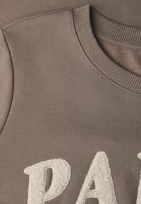 Brązowa bluza z bliska, z teksturowanym, kremowym haftowanym logo "P.A.R.I.S." na przodzie oraz prążkowanym dekoltem w stylu crewneck.