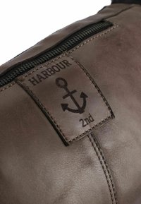 Borsa in pelle marrone con superficie testurizzata, dotata di una cerniera e di un'etichetta con un logo a forma di ancora e la scritta "HARBOUR 2nd" in rilievo.
