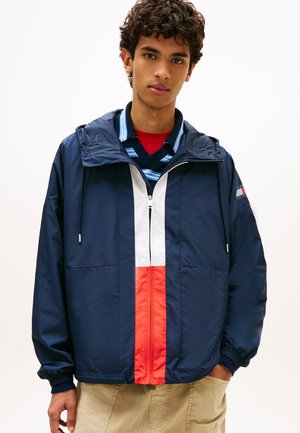 Jonge man die een marineblauwe windbreaker draagt met rood-witte verticale strepen, daaronder een gestreepte blauwe trui en beige broek.