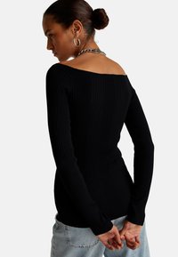 Top negro de manga larga acanalado con un diseño off-the-shoulder. Presenta una forma ajustada y un material suave y texturizado. Llevado con jeans de color azul claro.