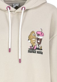 Beige Kapuzenpullover mit einem Frontdruck eines Croissants und eines Getränkewesens, ausgestattet mit weißen Kordeln und rosa Akzenten an den Ösen.