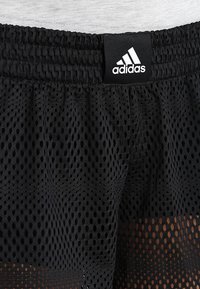 Pantalones cortos de malla negra con un patrón texturizado de grandes agujeros. Presentan una cintura elástica con una etiqueta del logotipo de Adidas en la parte frontal.