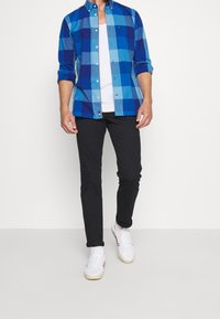 Hombre con una camisa de cuadros azul arremangada sobre una camiseta blanca, pantalones negros ajustados y zapatillas blancas de pie contra un fondo liso.