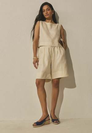 Žena s dlhými tmavými vlasmi, oblečená v bezrémom béžovom top a zodpovedajúcich šortkách po kolená, modré klinové sandále, stojaca opretá o štruktúrovanú stenu.